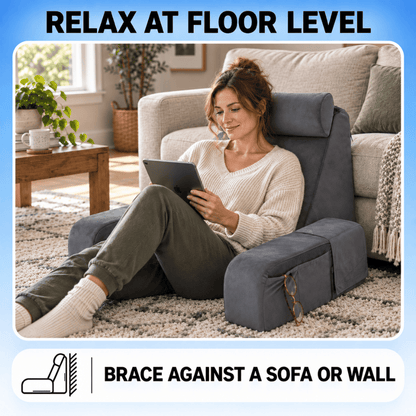 A woman sits on a dark gray floor-level reclining chair in a living room, holding a tablet. The chair is positioned in front of a sofa and wall, with text overlays reading 'RELAX AT FLOOR LEVEL' and 'BRACE AGAINST A SOFA OR WALL'.