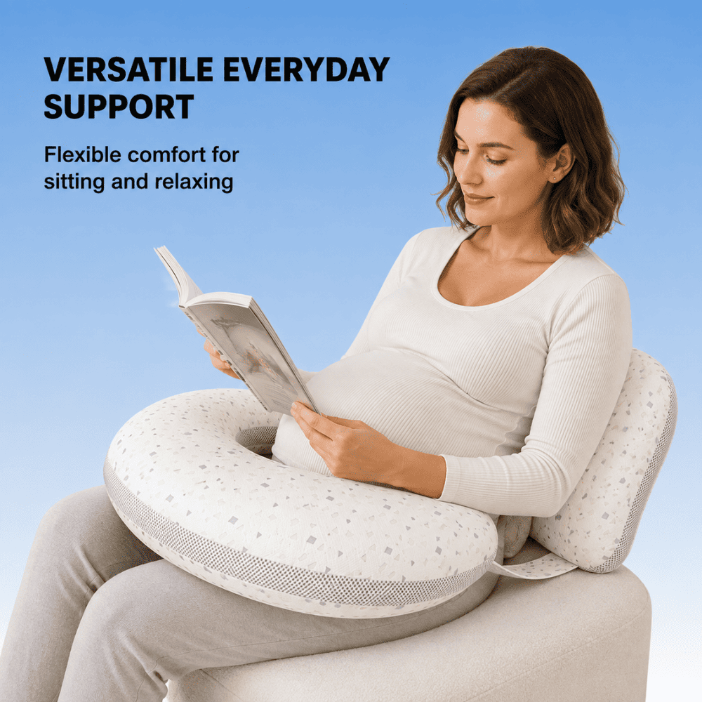 A pregnant woman sits on a cushioned support chair holding a book, wearing a white ribbed long-sleeve top and light gray pants. A white U-shaped nursing pillow with small gray dots rests around her belly.