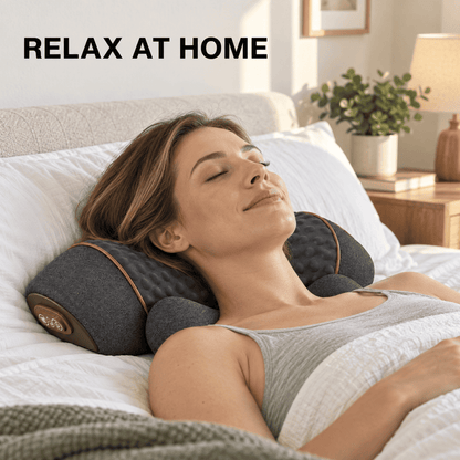 A woman lies relaxed on a bed using a dark gray contoured neck pillow with brown trim. The text 'RELAX AT HOME' appears in the top left corner, and a cozy bedroom setting with a lamp and plant is visible in the background.