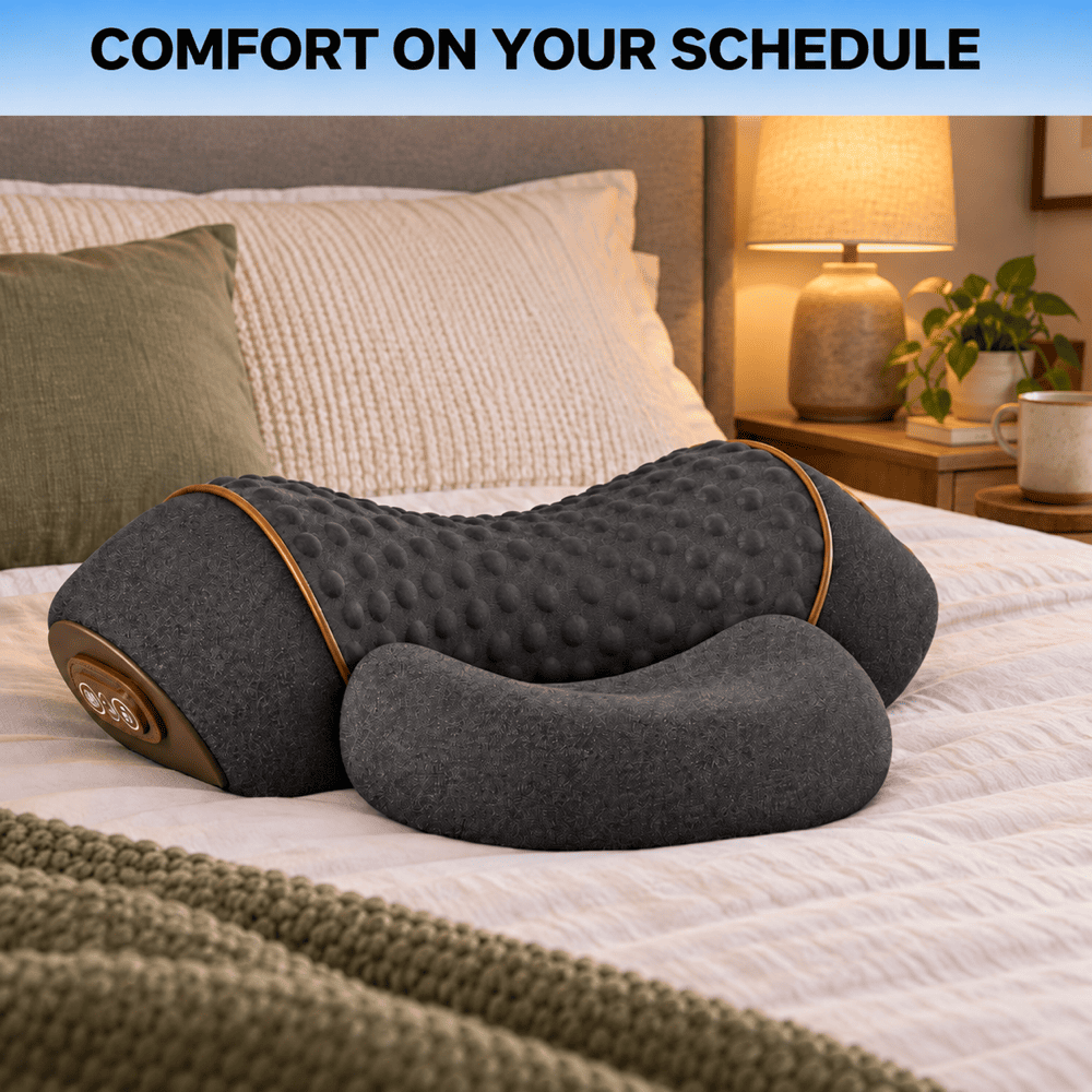 A dark gray contoured massage pillow with a textured dotted backrest and smooth head/foot sections, resting on a bed in a cozy bedroom setting. A text banner at the top reads 'COMFORT ON YOUR SCHEDULE'.