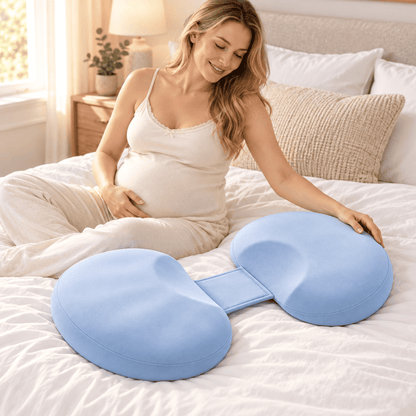A pregnant woman sits on a white bed using a light blue dual-sided maternity support pillow with a central connector, in a bright, warm-toned bedroom.